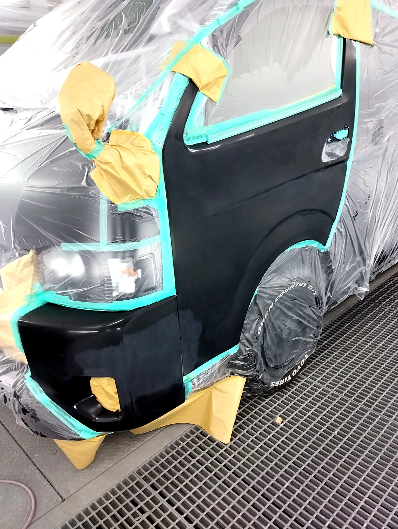 塗装ブースでビニールに覆われたハイエース。バンパー左角と左フロントドア1枚を出したところ（クリヤーの前）