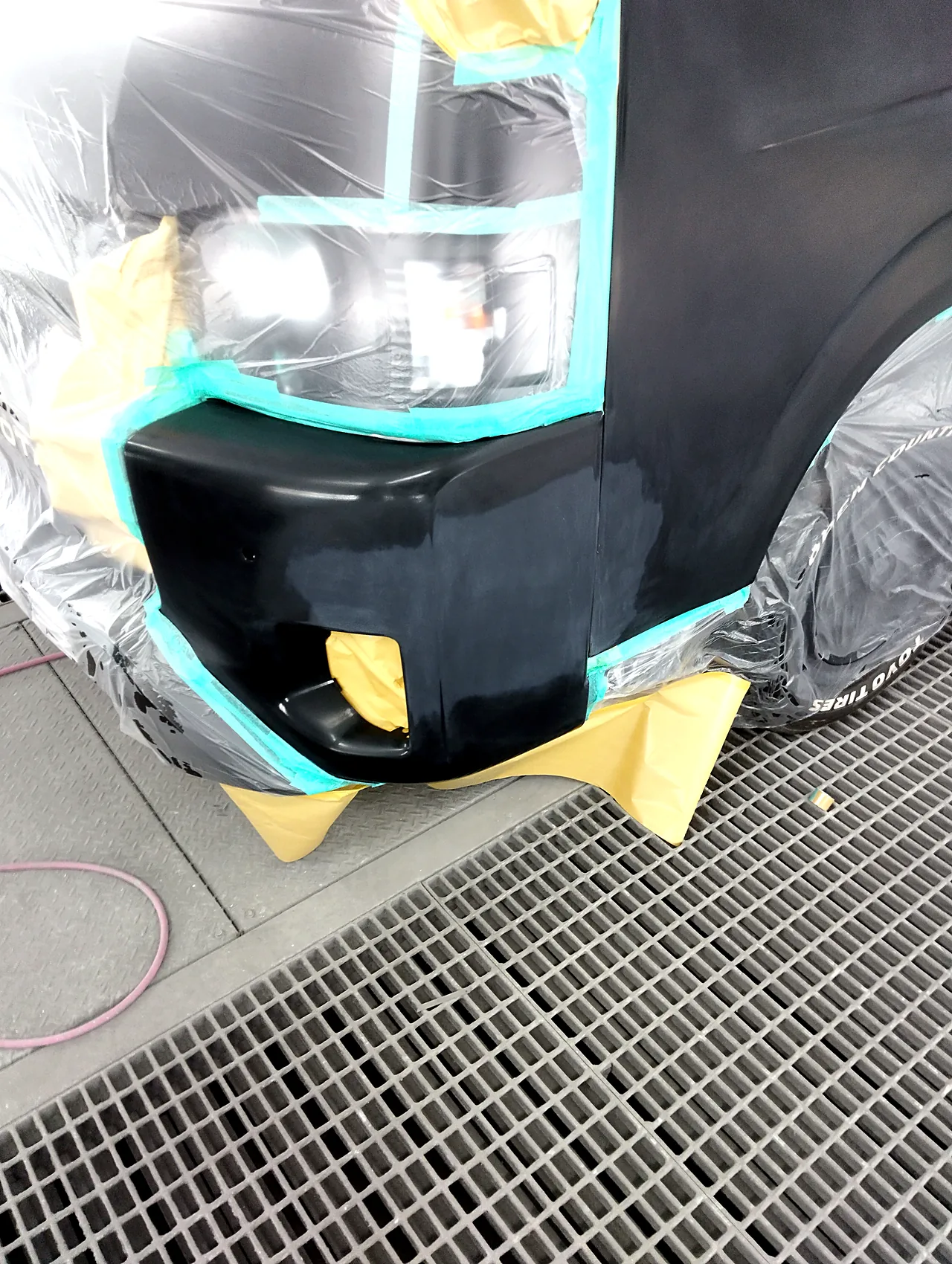 塗装ブースのハイエースのバンパー左角。ヘッドライトの縁にテープ、フォグランプの穴は裏から紙（クリヤーの前）