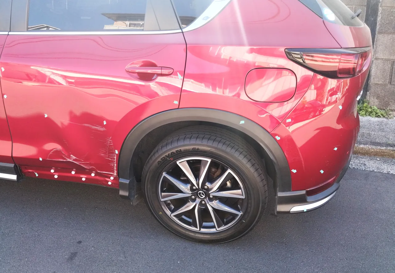 修理前のマツダ CX-5の左側面の後ろ側。左リヤドアの下半分のへこみと擦り傷、リヤフェンダーと後ろのバンパーにも目印のテープ
