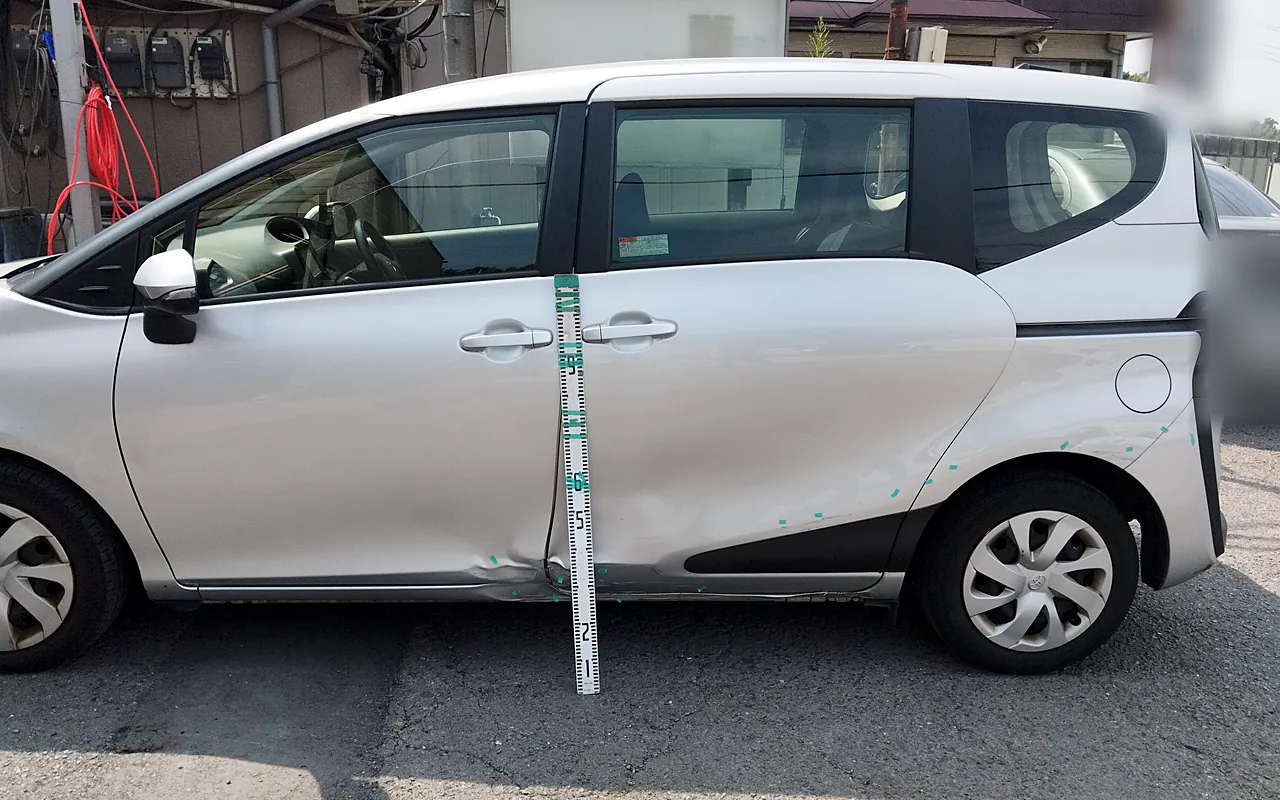修理の前のトヨタ シエンタの左側面を真横から。ドアの境目に目盛りの棒、スライドドアの下から後輪のまわりに緑のテープ