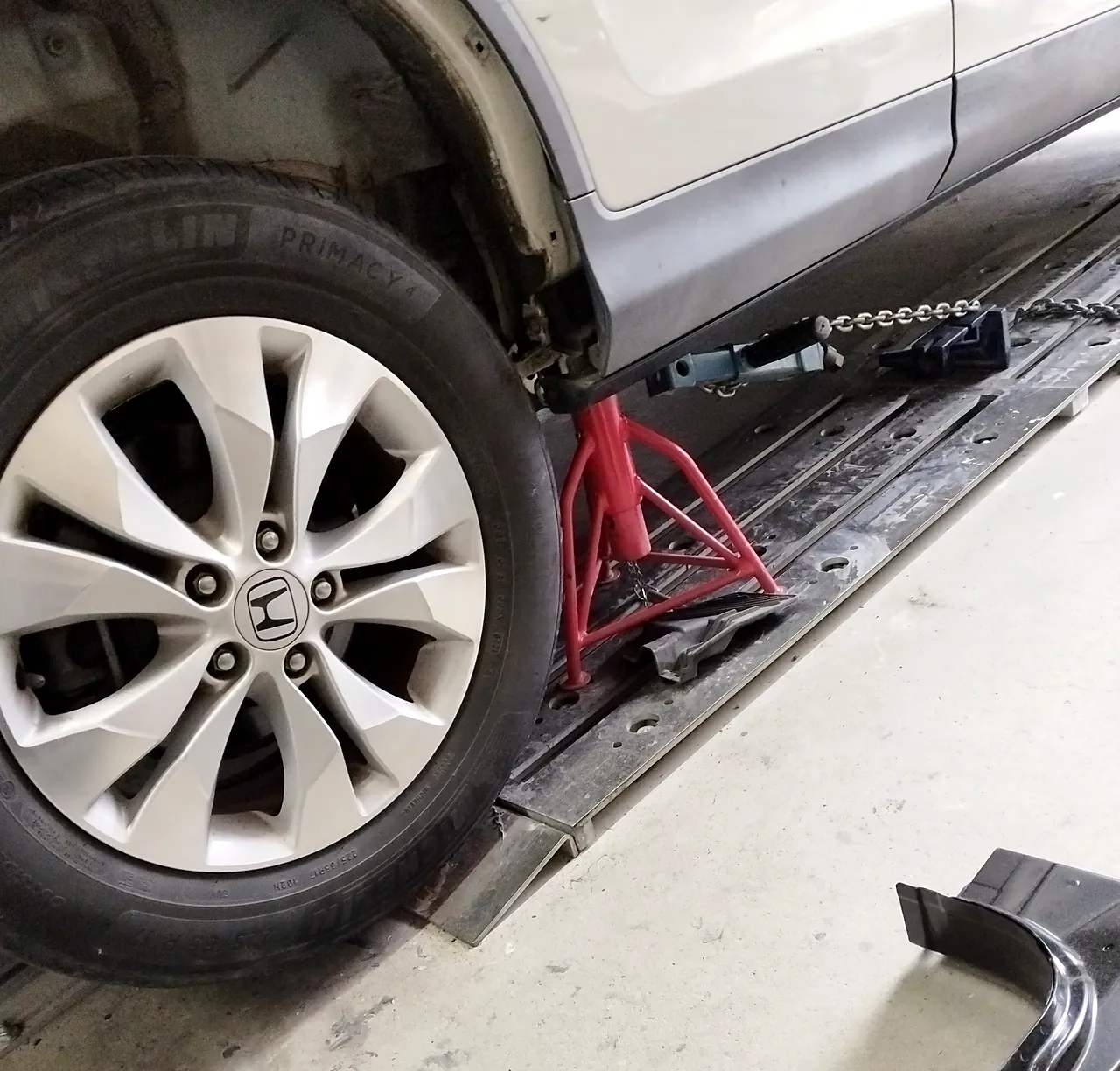 ホンダ CR-V の車体の下の縁（サイドシル）を赤いスタンドとクランプで留め、床のレールにチェーンでつなぐ