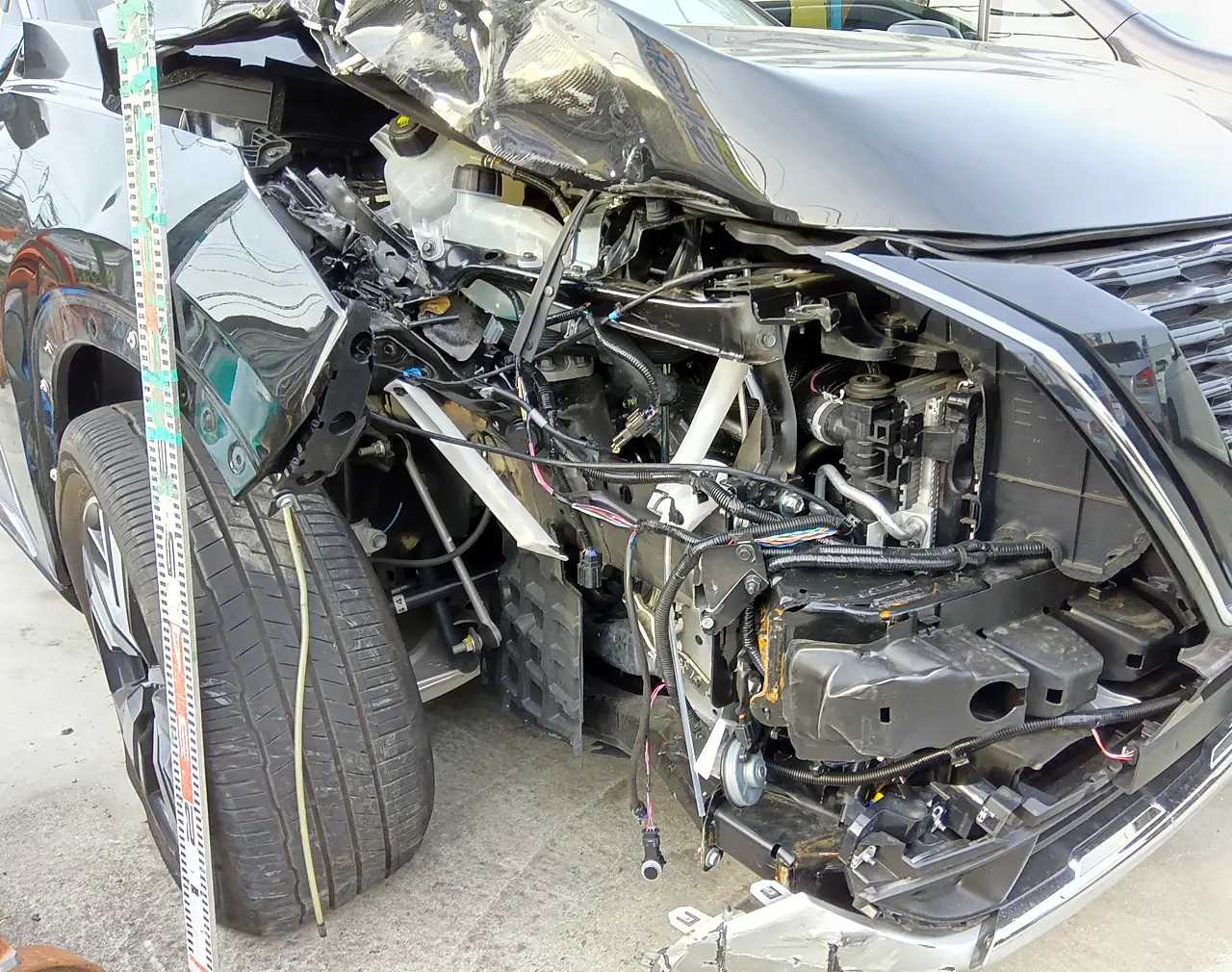 エクストレイル(T33)の右前の寄り。ヘッドランプがなく、配線や中の部品がむき出しで、右前輪が押し込まれている(修理前・目盛りの棒)